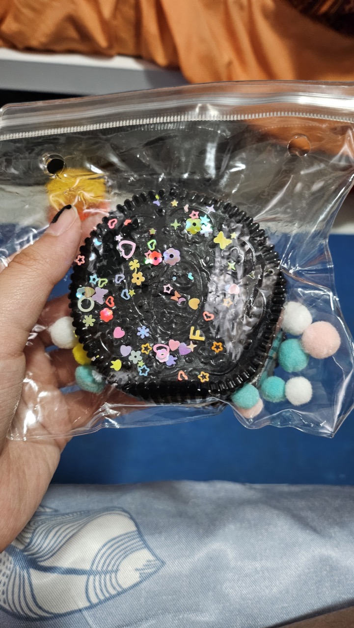 Picture of me holding up an Oreo-like taba squishy in a ziplock bag. It's covered in sequins and pom-poms.